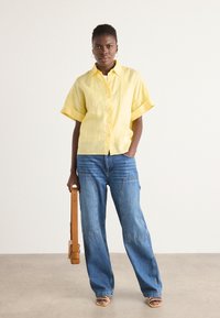 Camicia gialla di lino con maniche arrotolate, abbinata a jeans di denim larghi. Borsa a tracolla marrone e sandali con tacco beige.