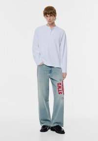 Vit långärmad henleytröja med tre knappar framtill, kombinerad med ljusblå vida jeans med röd "STWD"-text och stjärndekor.