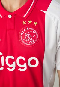 Maillot de football Ajax rouge et blanc, avec une texture en maille, col polo, logo du club avec un lion, et trois étoiles dorées au-dessus du logo.