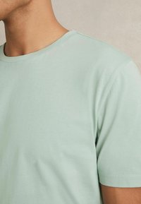 Tricou din bumbac verde deschis, cu guler rotund, mâneci scurte și textură netedă, prezentat din lateral, fără modele sau accente vizibile.