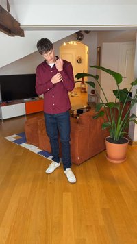 Hnedočervená košeľa na gombíky s dlhým rukávom, nosená s tmavými džínsami a bielymi teniskami, stojaca vedľa kvetináča v útulnom obývacom priestore.