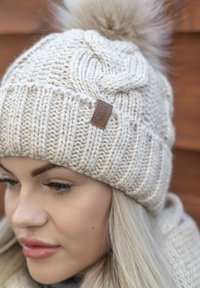 Gorro de punto en beige claro con un patrón texturizado, rematado con un pompón esponjoso y una pequeña etiqueta de cuero en el lado.