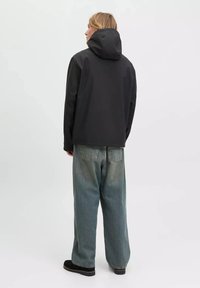 Person mit langen blonden Haaren, der eine schwarze Kapuzenjacke, lockere, ausgewaschene blaue Jeans und schwarze Schuhe trägt, steht mit dem Rücken zu einem schlichten Hintergrund.