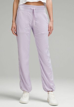 Femme portant un pantalon de jogging lilas à cordon avec poignets élastiques et des baskets blanches, debout devant un fond gris uni.