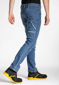 Jean en denim bleu à coupe slim, avec plusieurs poches et des coutures contrastantes, associés à des chaussures de sécurité noir et jaune.