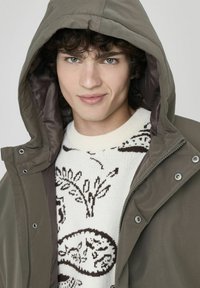 Parka à capuchon vert olive avec fermeture éclair et boutons-pression, superposée à un pull crème avec des motifs floraux bruns et des textures.