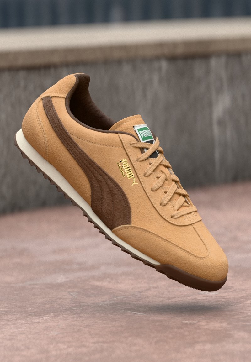 Sapatilha em camurça bege com detalhes castanhos, sola de borracha, atacadores planos e um rótulo verde. Apresenta um logotipo dourado na lateral. Design leve.