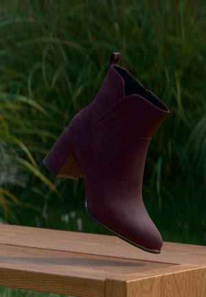 Bottine en daim violet foncé à talon carré lévitant au-dessus d'un banc en bois en extérieur avec un fond d'herbe haute.