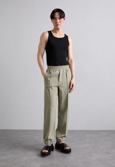 Musthave musta tops, heledad hallid hallidadega pleeded püksid elastse v�öökohtaga ja mustad platvormsandlid, seistes neutraalse tausta ees.