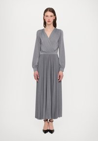 Robe grise plissée avec des manches longues et un design croisé, présentant une taille ajustée et une jupe fluide, fabriquée dans un tissu doux. Chaussures noires visibles.