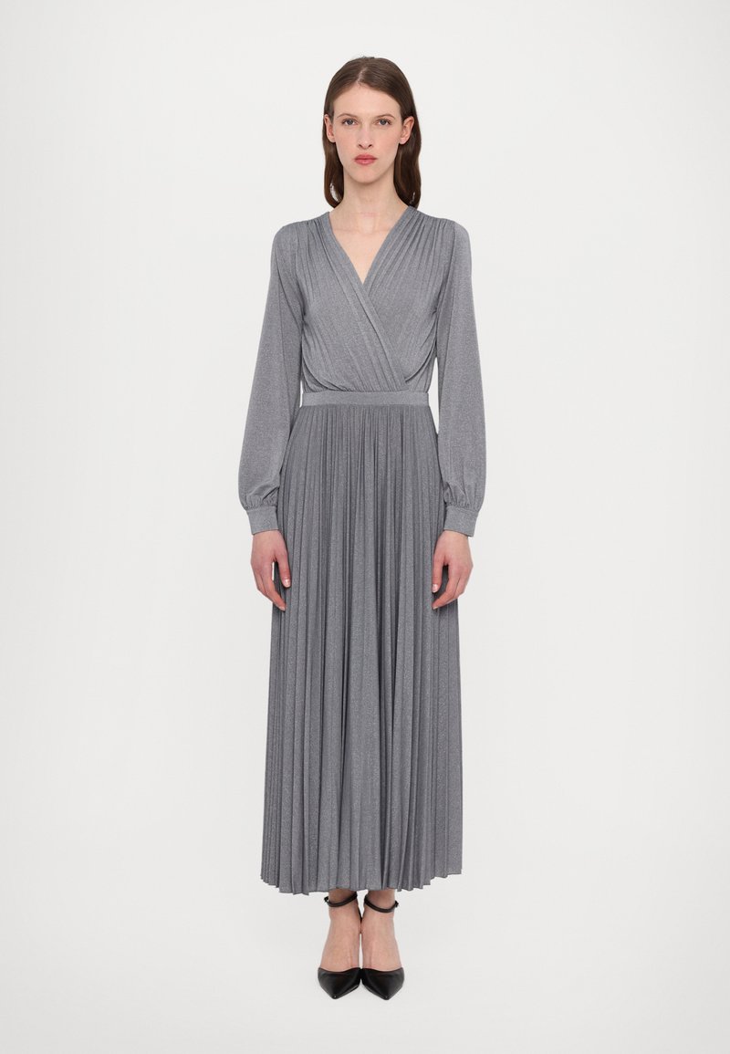 Robe grise plissée avec des manches longues et un design croisé, présentant une taille ajustée et une jupe fluide, fabriquée dans un tissu doux. Chaussures noires visibles.