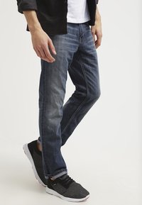 Vaqueros oscuros de hombre en denim, con corte recto, detalles de desvanecimiento y un estilo casual, combinados con zapatillas deportivas negras.