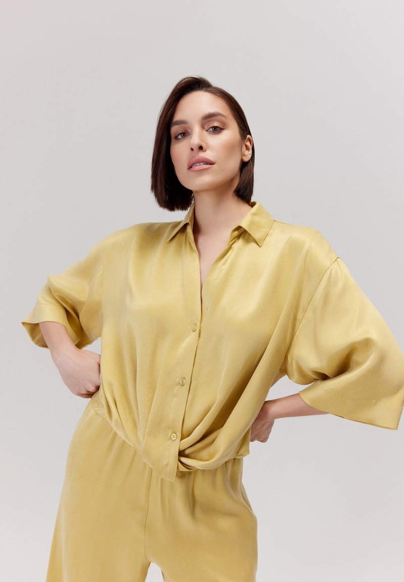 Żółta satynowa koszula o luźnym kroju, z zapięciem na guziki z przodu i krótkimi rękawami. Gładka tekstura, lekko marszczona w talii.