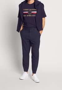 Námořnické tričko se zlatým textem a logem, spárované s námořnickými proužkovanými kalhotami a bílými teniskami. Svetr přehozený přes ramena.