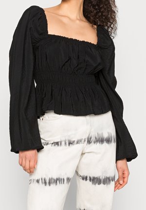 Woman wearing a black ruched long-sleeve blouse with puffed sleeves and white tie-dye jeans with black horizontal stripes.
