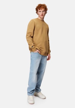 Khakifarbenes Sweatshirt mit Rundhalsausschnitt, gerippten Bündchen und einem Logopatch; kombiniert mit hellblauen, lockeren Jeans und weißen Sneakers.