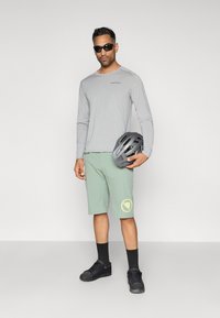 Svijetlosiva majica dugih rukava, zelene kratke hlače s perforacijama i žutim logotipom, crne sunčane naočale i tenisice, drži kacigu.