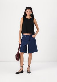 Svart kort topp, blå denimshorts med avslappnad passform och svarta flats. Modellen håller en vinröd handväska mot en enkel bakgrund.