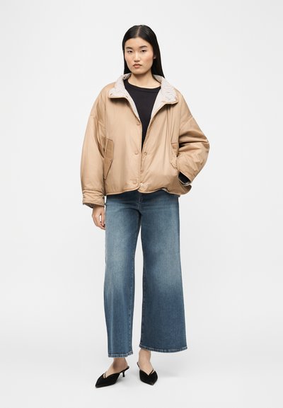 Γυναίκα που φορεί μπεζ μπουφάν με ριγέ γιακά, μαύρο τοπ, παντελόνι τζιν φαρδιάς γραμμής μπλε και μαύρα πέδιλα με τακούνι, στέκεται με το ένα χέρι στην τσέπη.