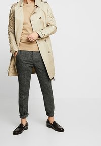 Manteau beige à double boutonnage, pull marron, pantalon texturé gris et mocassins en cuir noirs.