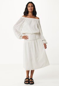 Vit off-shoulder klänning med broderade utskärningmönster, långa ballongärmar och cinchad midja, som matchas med tjocka svarta sandaler.