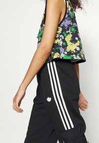 Ärmelloses Oberteil mit floralem Muster in Schwarz, Grün, Lila und Gelb; kombiniert mit schwarzen Hosen mit weißen Streifen und einem Logo-Detail.