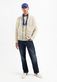 Man met blauwe pet, blauwe bandana om de nek, beige fleece vest, crèmekleurige trui, donkere spijkerbroek en witte sneakers staande tegen een witte achtergrond.