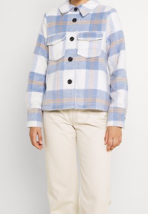 Femme portant une veste boutonnée à carreaux bleus, blancs et beiges avec des poches poitrine à rabat, et un pantalon couleur crème.