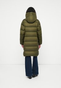 Abrigo acolchado verde oliva con capucha, que presenta paneles acolchados horizontales, corte de longitud media y bolsillos laterales. Llevado sobre jeans oscuros de campana.