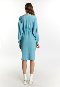 Šviesiai mėlyna, tekstūruota suknelė su ilgomis rankovėmis, susiaurinta juosmenyje, iki kelių ilgio ir blizgiu finišu. Apsirengta su baltais kulkšninių botais.