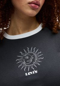 Κοντινό πλάνο ενός ατόμου με σγουρά μαλλιά που φοράει μαύρο μπλουζάκι Levi's με λευκό σχέδιο ήλιου και φεγγαριού στο στήθος.