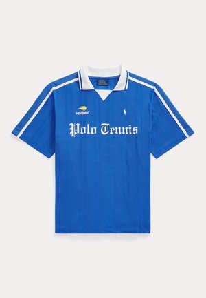 Homme debout portant un polo de tennis bleu, un pantalon bleu marine et des baskets blanches sur un fond blanc uni.