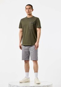 Oliven grønn bomulls t-skjorte med brystlomme, kombinert med lys grå shorts og hvite sneakers, stående på en marmoroverflate.