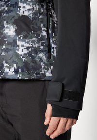 Jachetă multicoloră cu un model de camuflaj în nuanțe de gri și negru, cu o textură netedă, manșetă cu Velcro ajustabil și buzunar cu fermoar.