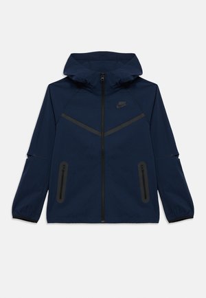 Casaco Nike azul-marinho com fecho de correr frontal, capuz e punhos elásticos. Apresenta detalhes em preto e dois bolsos laterais com fecho de correr. Textura lisa.