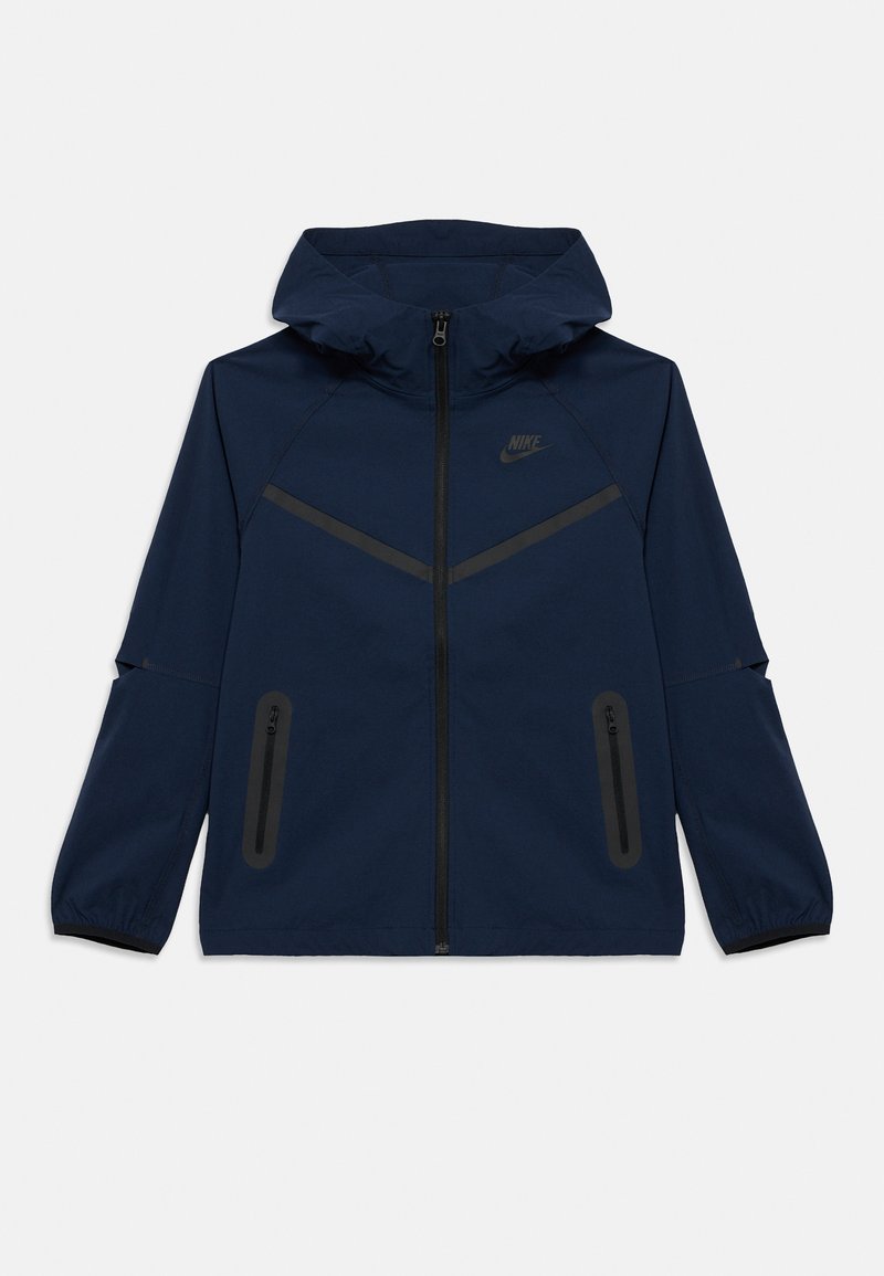 Casaco Nike azul-marinho com fecho de correr frontal, capuz e punhos elásticos. Apresenta detalhes em preto e dois bolsos laterais com fecho de correr. Textura lisa.