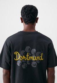 Svart t-skjorte med teksturert, stilisert gull "Dortmund"-tekst mot en subtil grå poter bakgrunn. Korte ermer, løs passform.