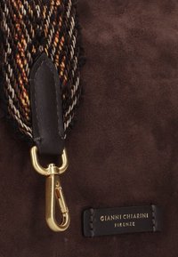 Dettaglio di una borsa in suede marrone con una tracolla intrecciata in tonalità calde, un fermaglio in oro e un'etichetta in pelle nera con la scritta "GIANNI CHIARINI FIRME" impressa.