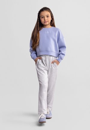 Jeune fille aux longs cheveux portant un sweat-shirt lavande, un pantalon de survêtement gris clair et des baskets violettes, debout les mains dans les poches sur un fond gris.