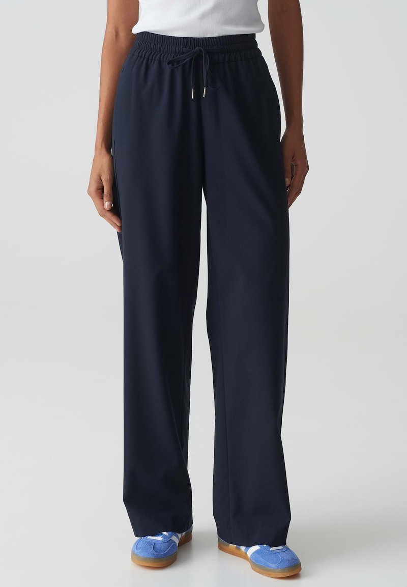 Pantalons bleu marine à taille élastique et jambes larges, avec cordon de serrage. Fabriqués en tissu doux, présentant une texture lisse et un minimum de détails.