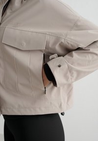 Licht beige jas met klepzak en drukknoopmanchet, hand in zijritszak gestoken, gedragen over zwarte geribde broek.