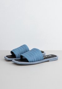 Paired de sandales à enfiler tissées bleu clair avec semelles intérieures noires et semelles plates sur une surface grise avec un fond blanc.