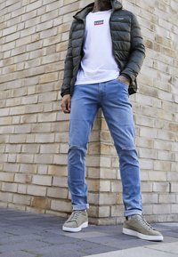 Chaqueta verde acolchada, camiseta blanca con logo rojo y azul, jeans de mezclilla azul claro con rodillas rasgadas y zapatillas gris-verdosas apoyadas contra una pared de ladrillo.