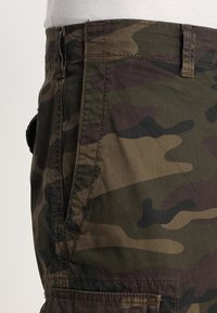 Detailní záběr na zelené a hnědé maskáčové cargo šortky, které jsou kombinovány s bílým tričkem, ukazující postranní kapsu a poutka na opasek.