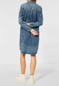 Person som står med armarna korsade bakom ryggen, iklädd en knälång blå denimklänning och vita högstövlade sneakers på en neutral bakgrund.