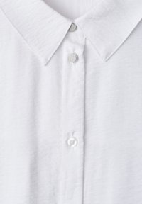 Chemise blanche à col avec une patte comportant un bouton magnétique argenté en haut et des boutons blancs classiques en dessous.