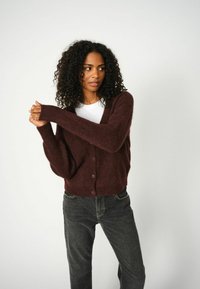 Cardigan bordeaux in tessuto morbido con scollo a V, maniche lunghe e cinque bottoni anteriori, abbinato a jeans scuri.