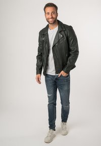 Giacca da biker in pelle verde scuro con tasche con zip, abbinata a una maglietta grigia, jeans blu strappati e sneaker bianche.