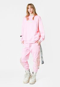 Sudadera rosa y joggers a juego hechos de material suave; cuenta con un parche gráfico, un patrón de plumas en los pantalones y correas de acento grises.