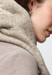 Écharpe en tricot beige drapée autour du cou, présentant une texture côtelée. L'arrière-plan est uni, mettant en valeur les détails et la douceur de l'écharpe.